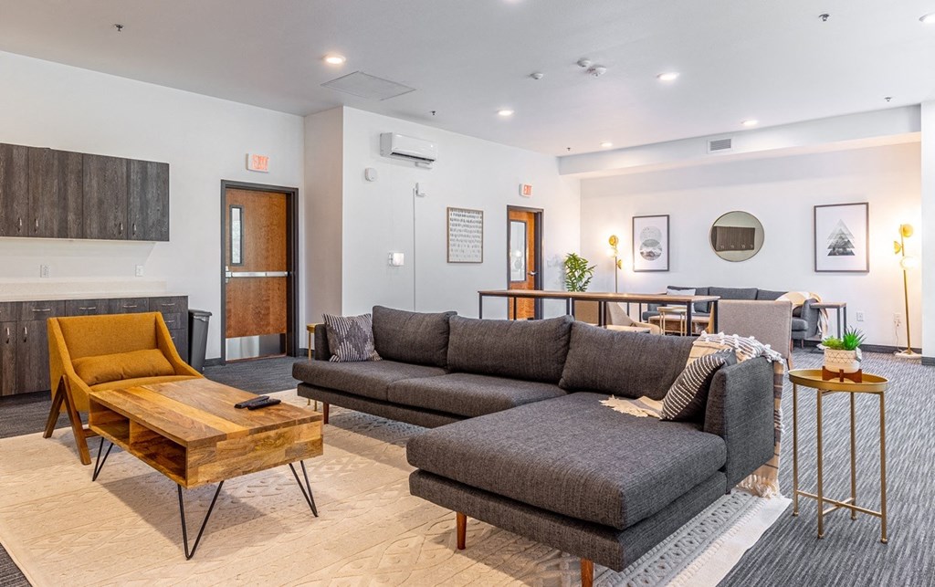 Vancouver, WA Apartments - 134th Street Lofts - Resident Lounge with Carpeted Flooring, Sectional Couch, Armchair, Wooden Coffee Table, Table with Chairs, and Wood-Tone Cabinetry.