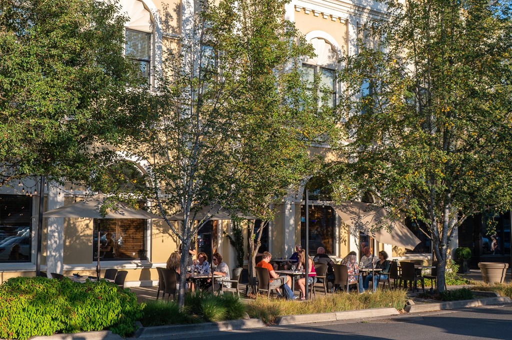 A group of people are sitting at tables under trees outside a building.
