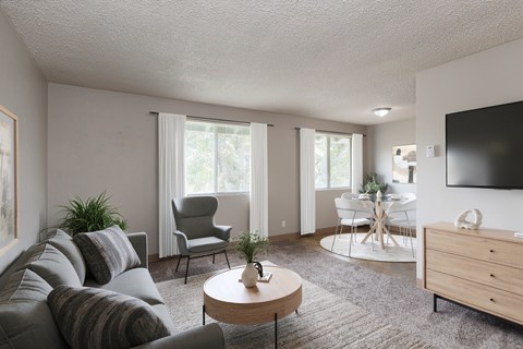 A living room with a grey couch, a wooden coffee table, a grey armchair, a dining table with chairs, and a flat-screen TV mounted on the wall.
