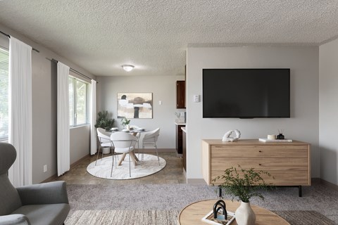 A modern living room with a grey sofa, a white dining table and chairs, a wooden cabinet, and a flat-screen TV mounted on the wall.