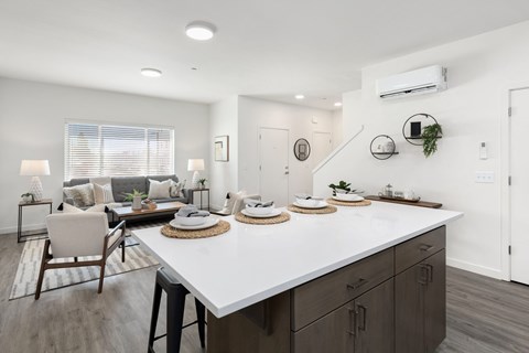 A modern kitchen with a white countertop and dark wood cabinets open to the living room with a couch and two sitting chairs.