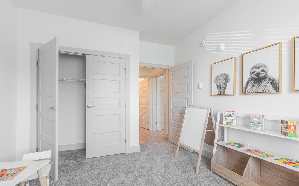 A room with a white door, a white table, a white chair, a white shelf with books and a white wall with a picture of a sloth and an elephant.