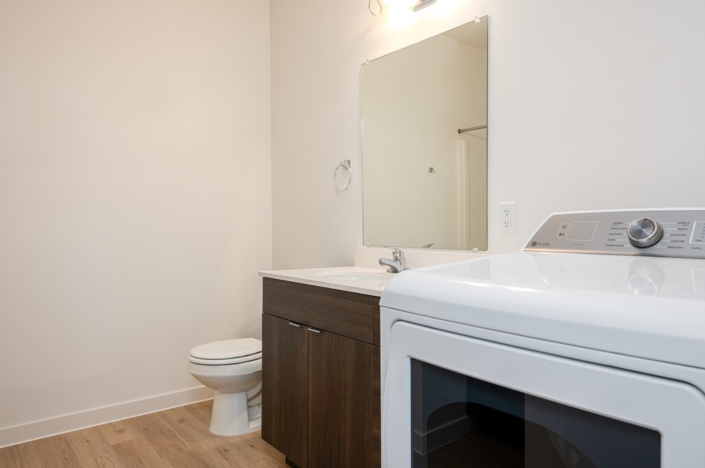 A white washing machine sits in a small laundry room with a toilet and a mirror.