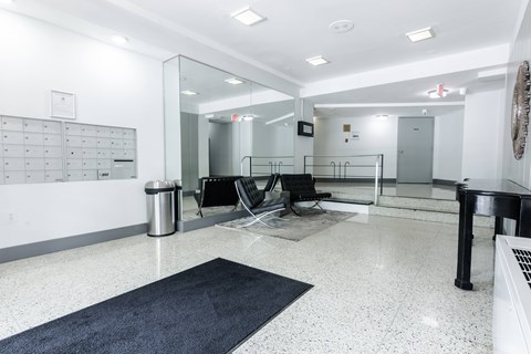 A hallway with a black and white tiled floor, a black trash can, a black chair, a black table, and a white wall with a calendar on it.