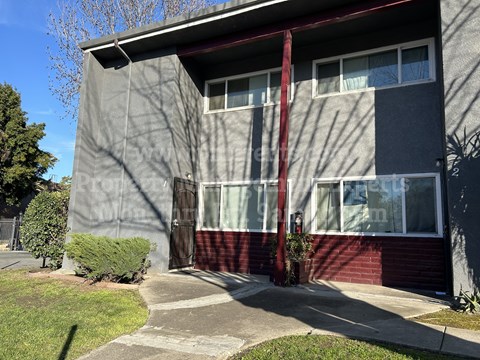 A grey and red building with a red awning and a small tree in front.