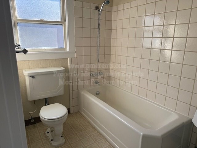 A white toilet is in a small bathroom with a white tub and a window.