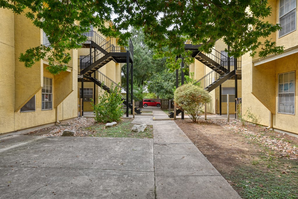 a sidewalk between two yellow buildings with trees and a red car at Mayfield Gardens, San Antonio, TX, 78211