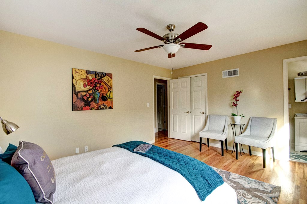 A bedroom with a bed, two chairs, a ceiling fan, and a painting on the wall.