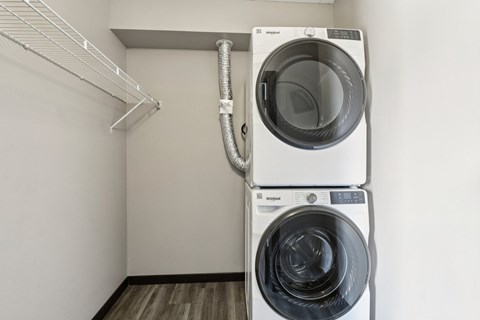 A white washing machine is in a small room with a white wall and a white shelf.