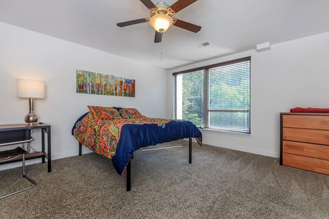 A bedroom with a bed, a desk, a chair, a ceiling fan, and a window with blinds.