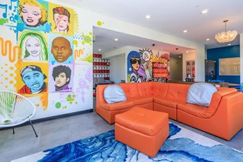 A brightly colored living room with a large orange sofa and a mural of various faces on the wall.