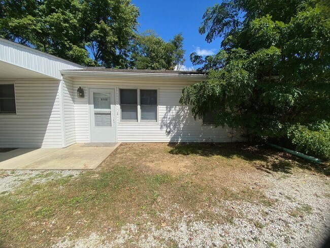 A small white house with a grey door and windows surrounded by trees.