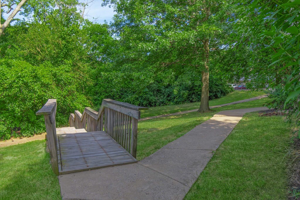 A wooden bridge over a concrete walkway surrounded by greenery.
