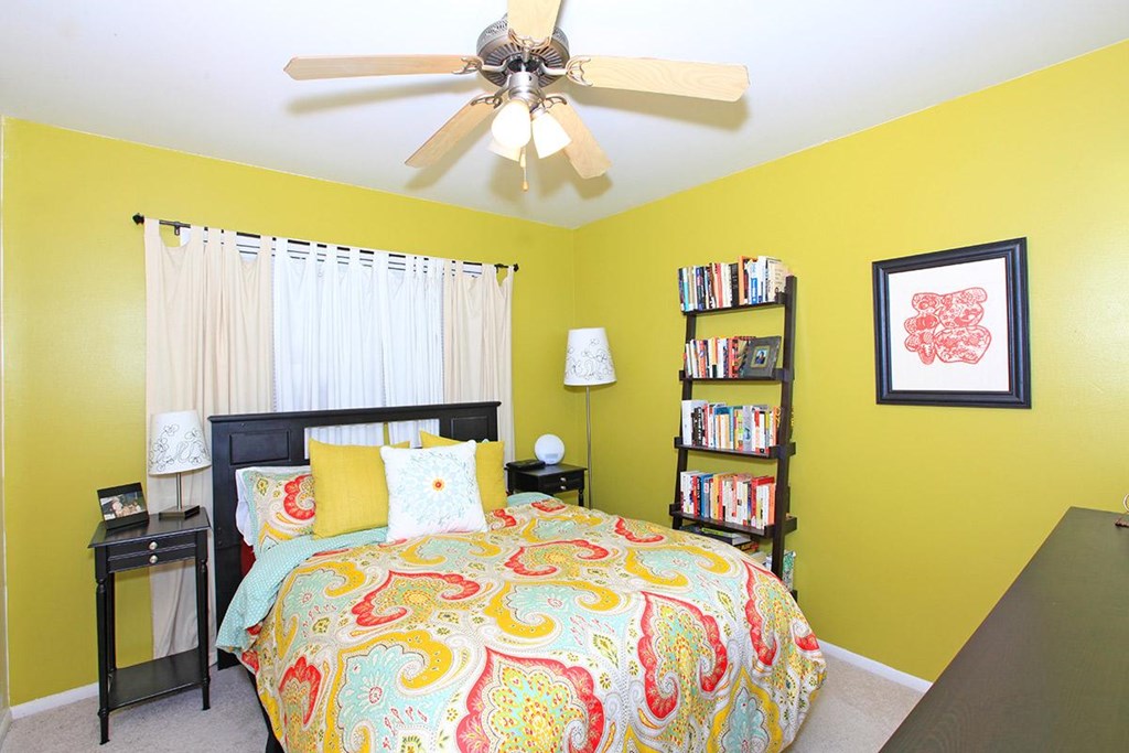 A bedroom with a bed, a ceiling fan, a nightstand, a lamp, a bookshelf, and a picture on the wall.