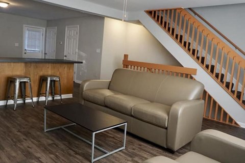 A brown couch sits in a living room with a bar and stairs in the background.