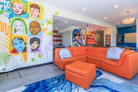 A brightly colored living room with a large orange sofa and a mural of various faces on the wall.