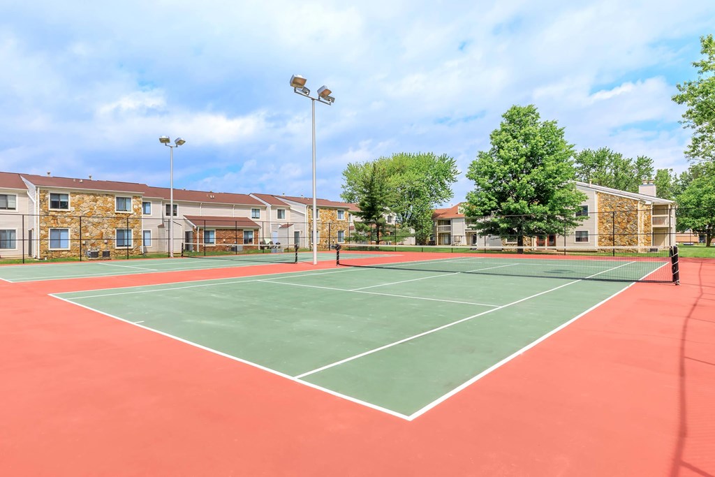 A tennis court with a red surface and white lines is surrounded by apartment buildings.