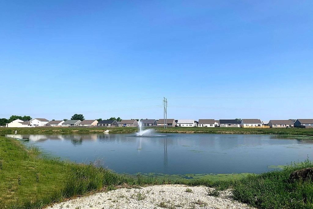 A serene lake with a fountain in the middle, surrounded by houses and a clear blue sky.
