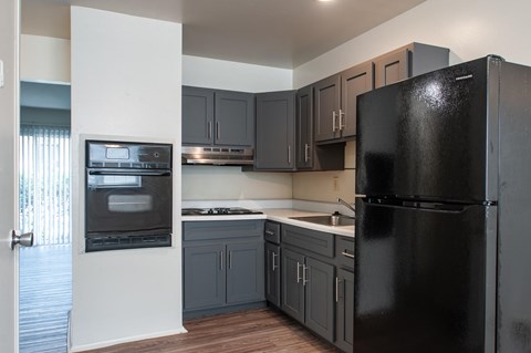 A black refrigerator in a kitchen with wooden floors and grey cabinets.