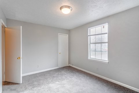 A room with a white door, a window with blinds, and a carpeted floor.