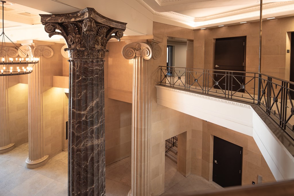 A large marble column stands in a spacious room with a balcony and a chandelier.