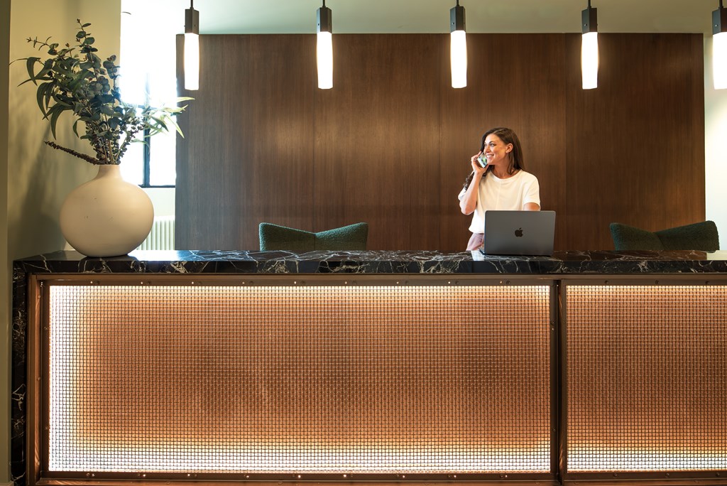 A woman is standing behind a reception desk with a laptop and answering a phone call.