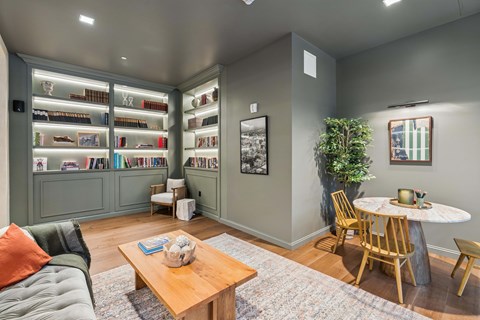 A living room with a grey couch, a wooden coffee table, a round table with chairs, and a bookshelf filled with books.