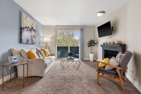 A living room with a grey sofa, a yellow cushion, a grey armchair, a glass table, a fireplace, a television, a plant, and a painting on the wall.