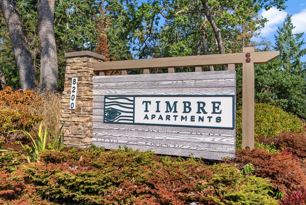 A sign for Timbre Apartments is displayed in front of a lush green background.