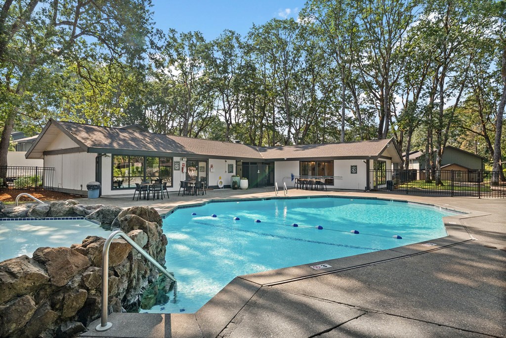 A large outdoor swimming pool surrounded by a stone wall and a patio area with chairs and a building in the background.