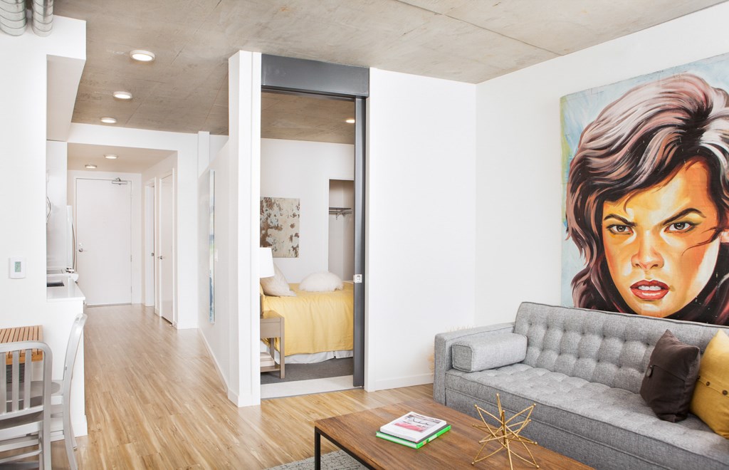 A living room with a grey couch and a painting of a woman's face on the wall.