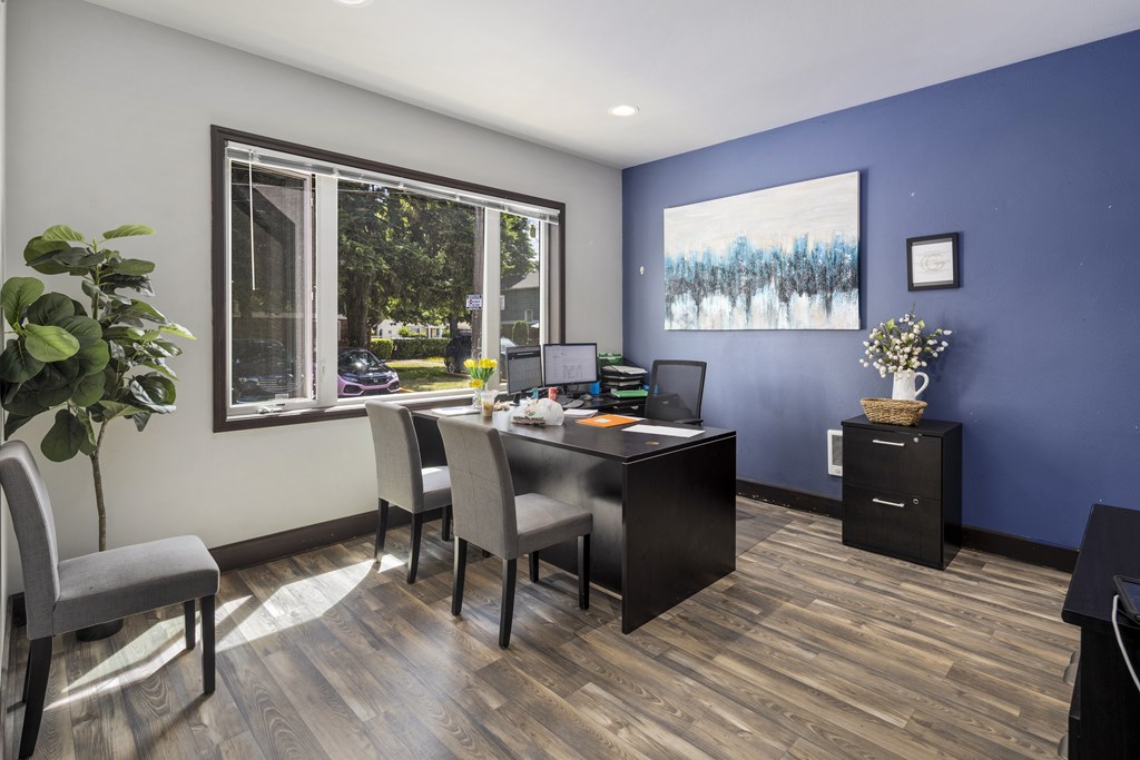 A room with a desk and chairs with a window and a painting on the wall.