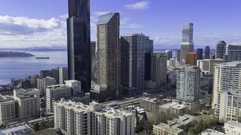 A cityscape with a mix of modern and older buildings, a body of water in the background, and a partly cloudy sky.