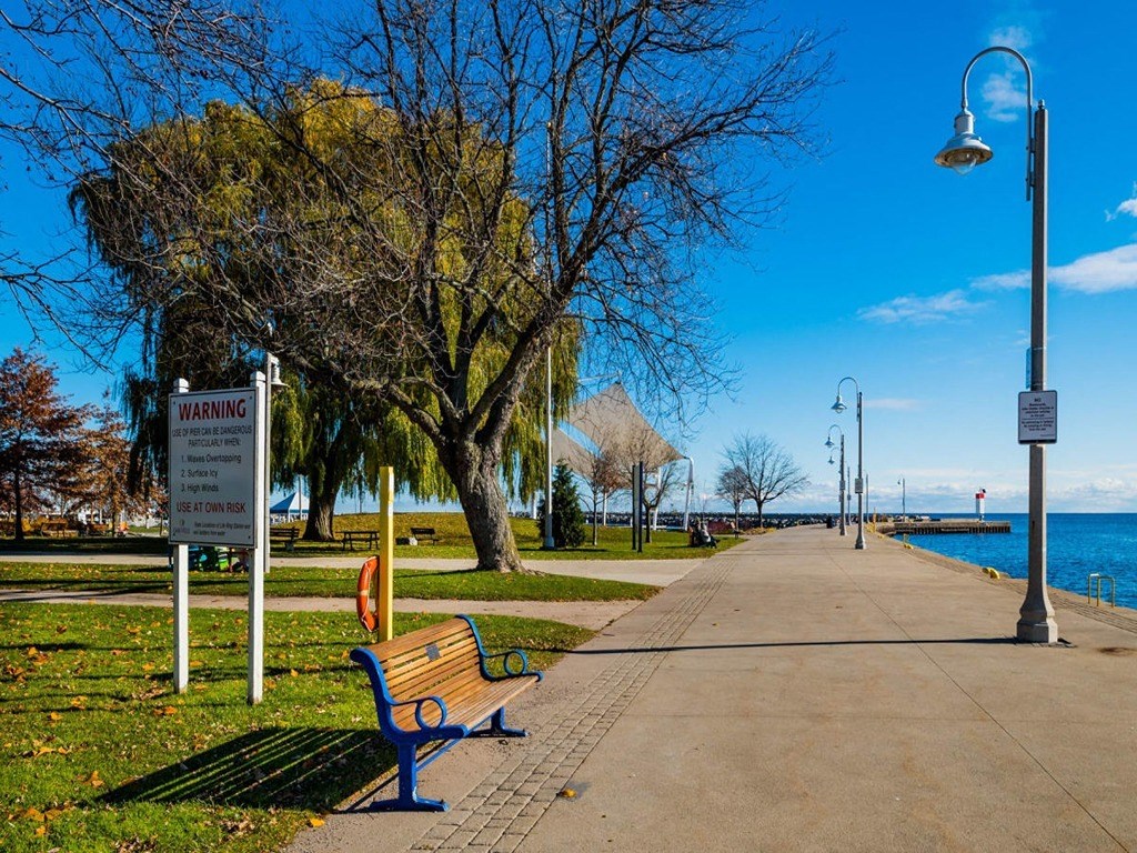 a park bench next to a sidewalk near the water  at The Village, Oakville, ON