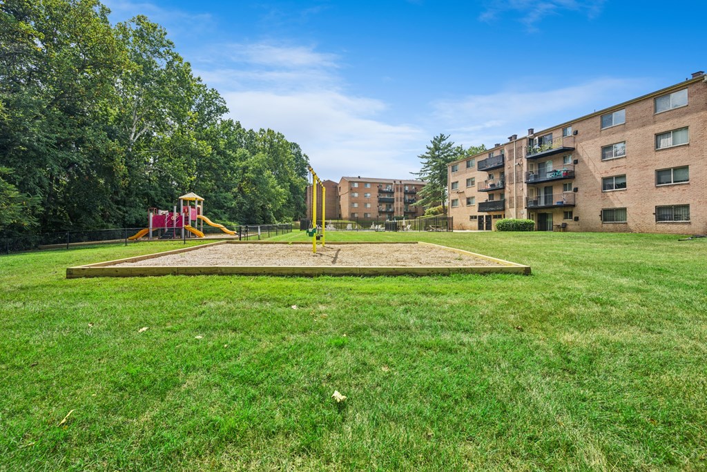 A playground with a slide and a yellow structure is in the foreground of a grassy area with apartment buildings in the background.