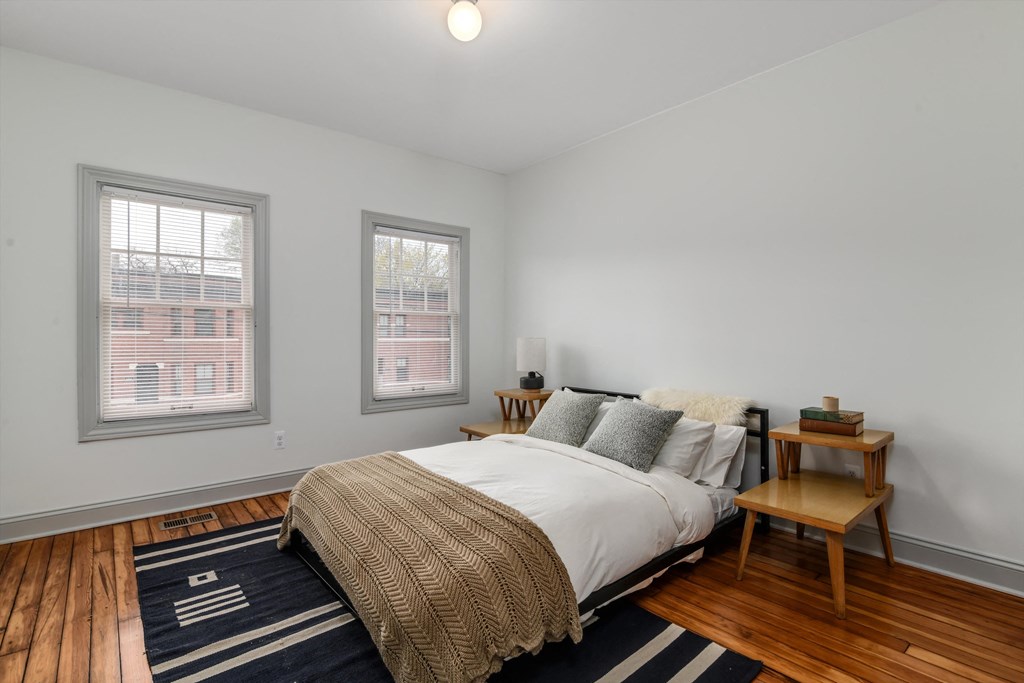 A bedroom with a bed, a small table, and two windows.
