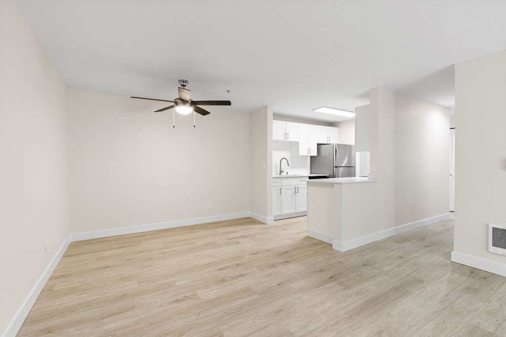 A room with a ceiling fan and light fixture, and a kitchen area in the background.