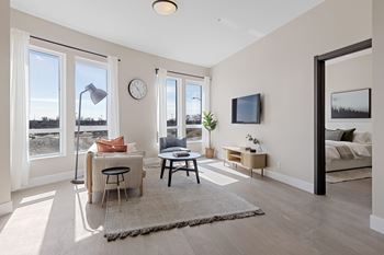 A living room with a sofa, a table, a lamp, a clock, a TV, and a window overlooking a field at The Woods at 180 Kanata Avenue, Kanata, ON