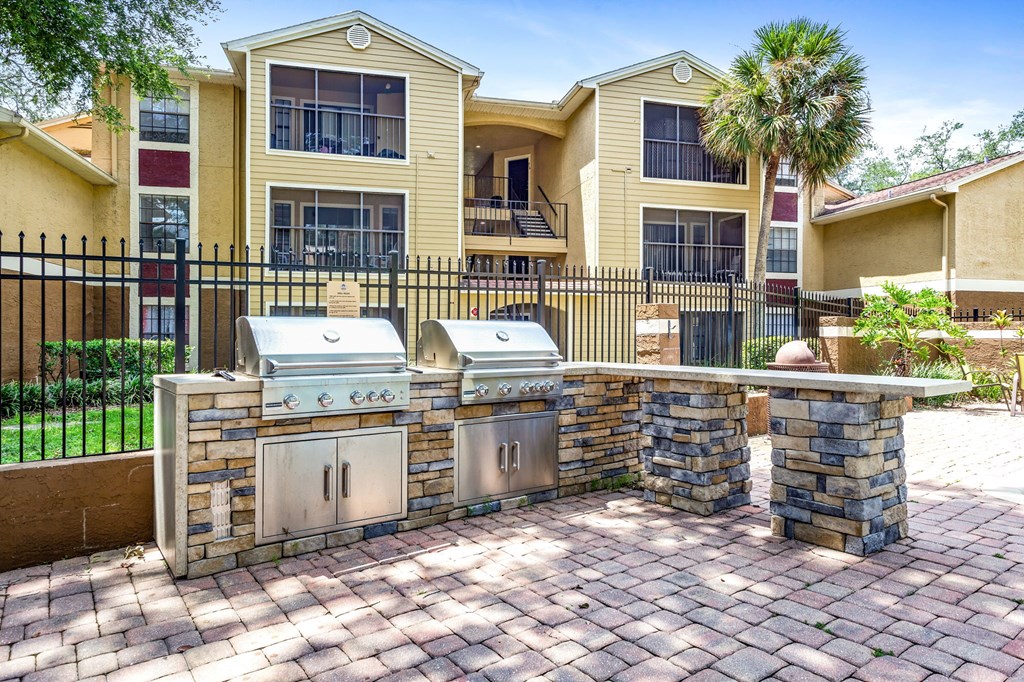A brick patio with a grill and a stone wall in front of a two-story house.