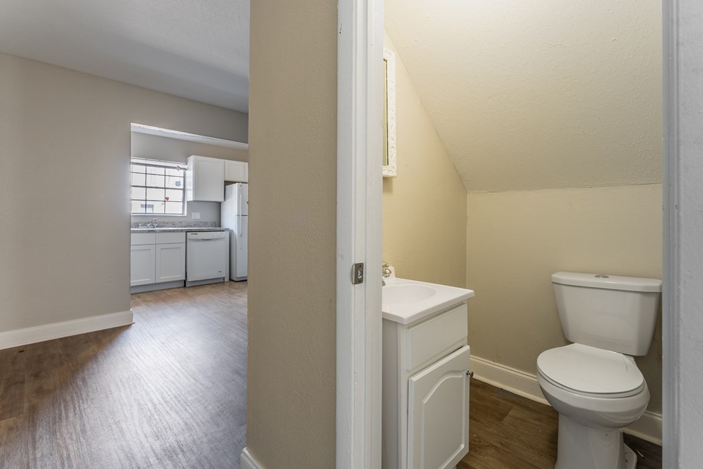 A white toilet sits in a bathroom next to a kitchen.