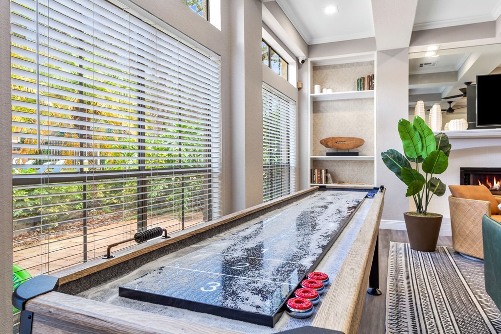 A pool table with red balls is in the foreground of a room with a window covered in blinds.