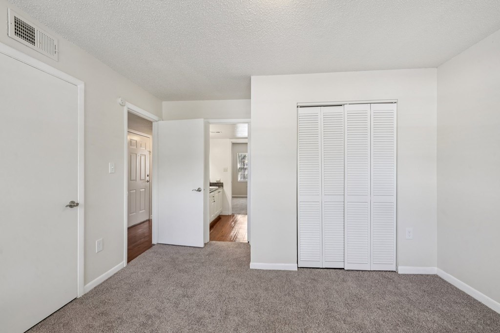 A room with white walls and carpeted floor with a white door on the left and a white closet with blinds on the right.