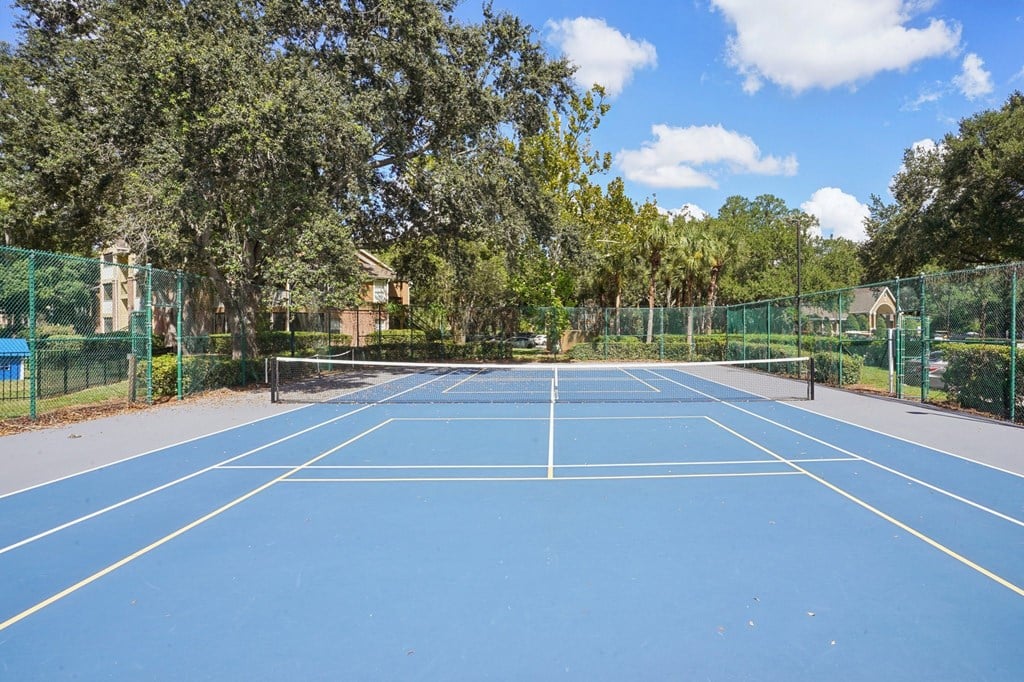 A blue tennis court surrounded by green fencing and trees.