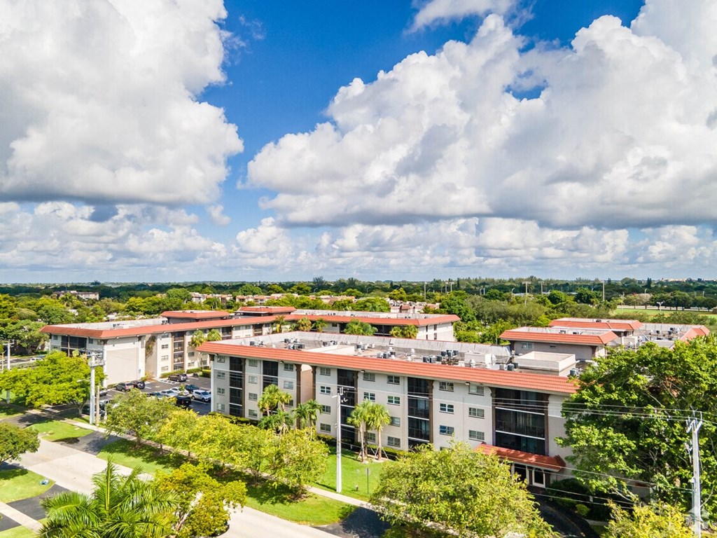 A large building complex with a red roof and white walls is surrounded by trees and has a grassy area in front.