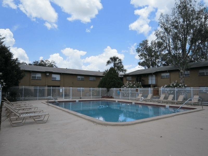 A pool surrounded by chairs and a fence in front of apartment buildings.