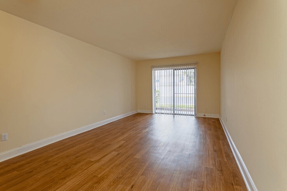 Large living room with warm toned wooden flooring and a sliding glass door with hanging blinds.