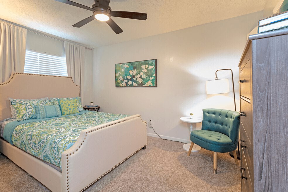 A spacious bedroom with light beige carpeting.. Double paned window with blinds.  Sleek ceiling fan.
