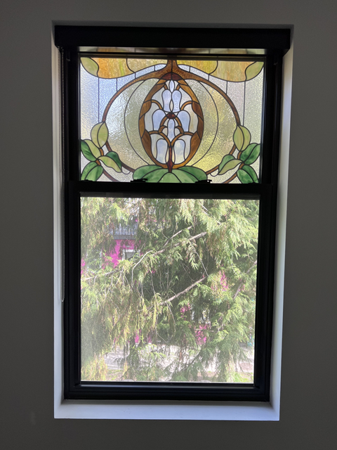 A stained glass window with a floral design at the top and a view of trees and a pink building outside.