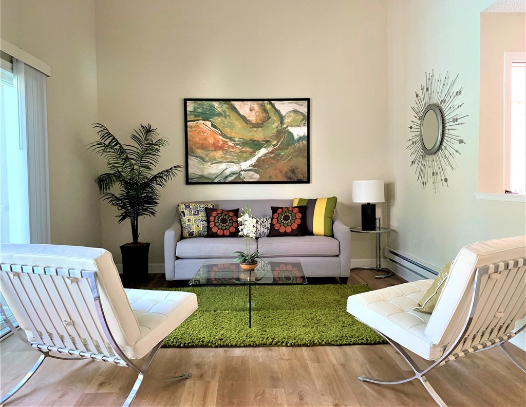 2 bedroom model living room with a tan couch, 2 white accent chairs, glass coffee table, green accent rug, plants, lamp and wall decorations
