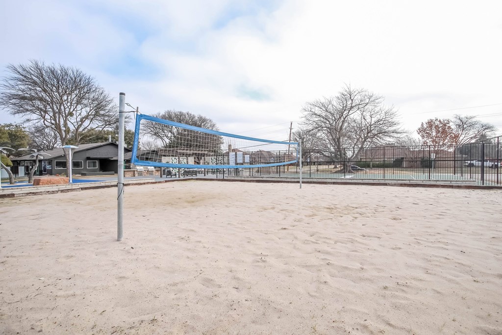 A sandy beach volleyball court with a blue net and a fence in the background.